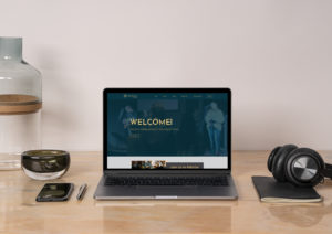 open laptop, iphone, and headphones displayed on a wood desk with a white wall in the background