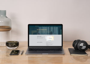 open laptop, iphone, and headphones displayed on a wood desk with a white wall in the background